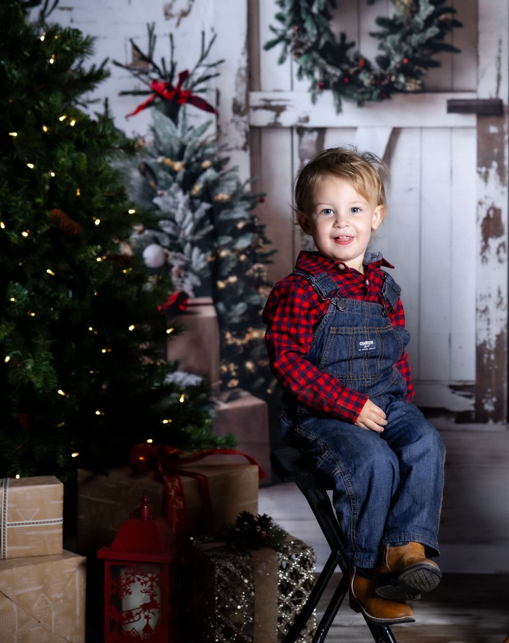 Small child in plaid shirt and overalls sits by decorated Christmas tree with wrapped presents.