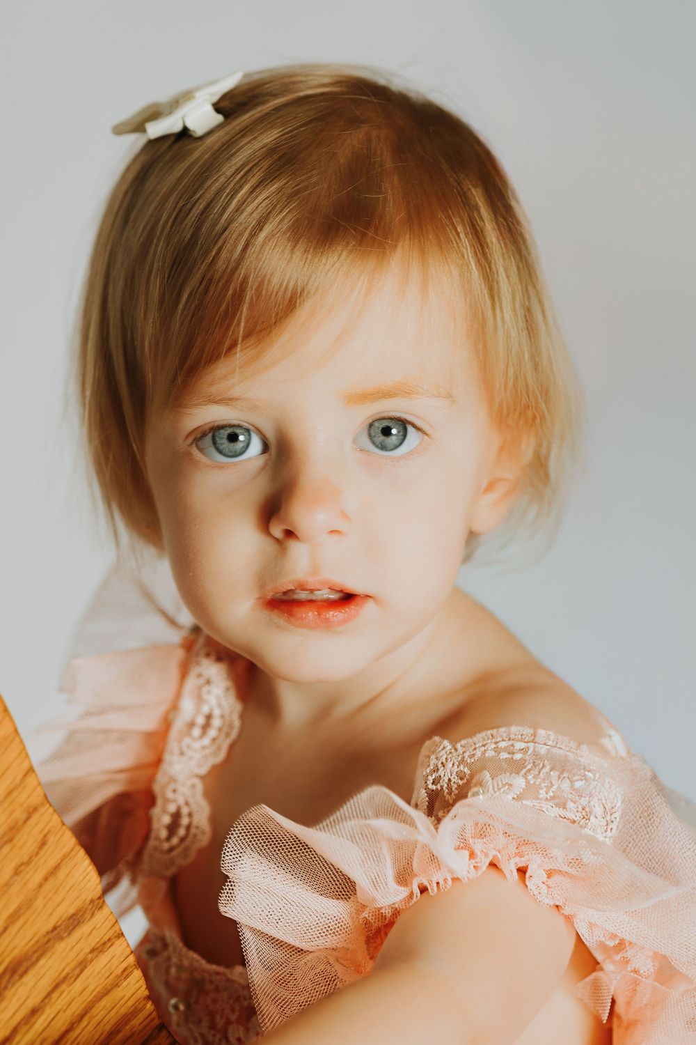 A child with bright blue eyes wears a delicate peach-colored lace dress and a small white hair clip.
