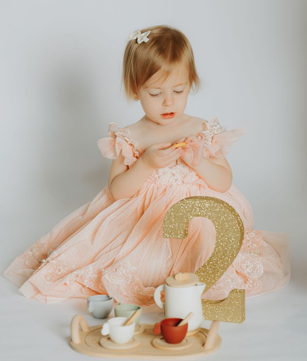 A toddler in a peach-colored dress sits next to a gold glitter number 2 and tea party setup on white background.