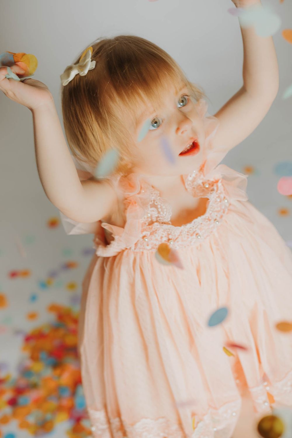 A small child in a peachy-pink dress dances joyfully amidst falling autumn-colored confetti.