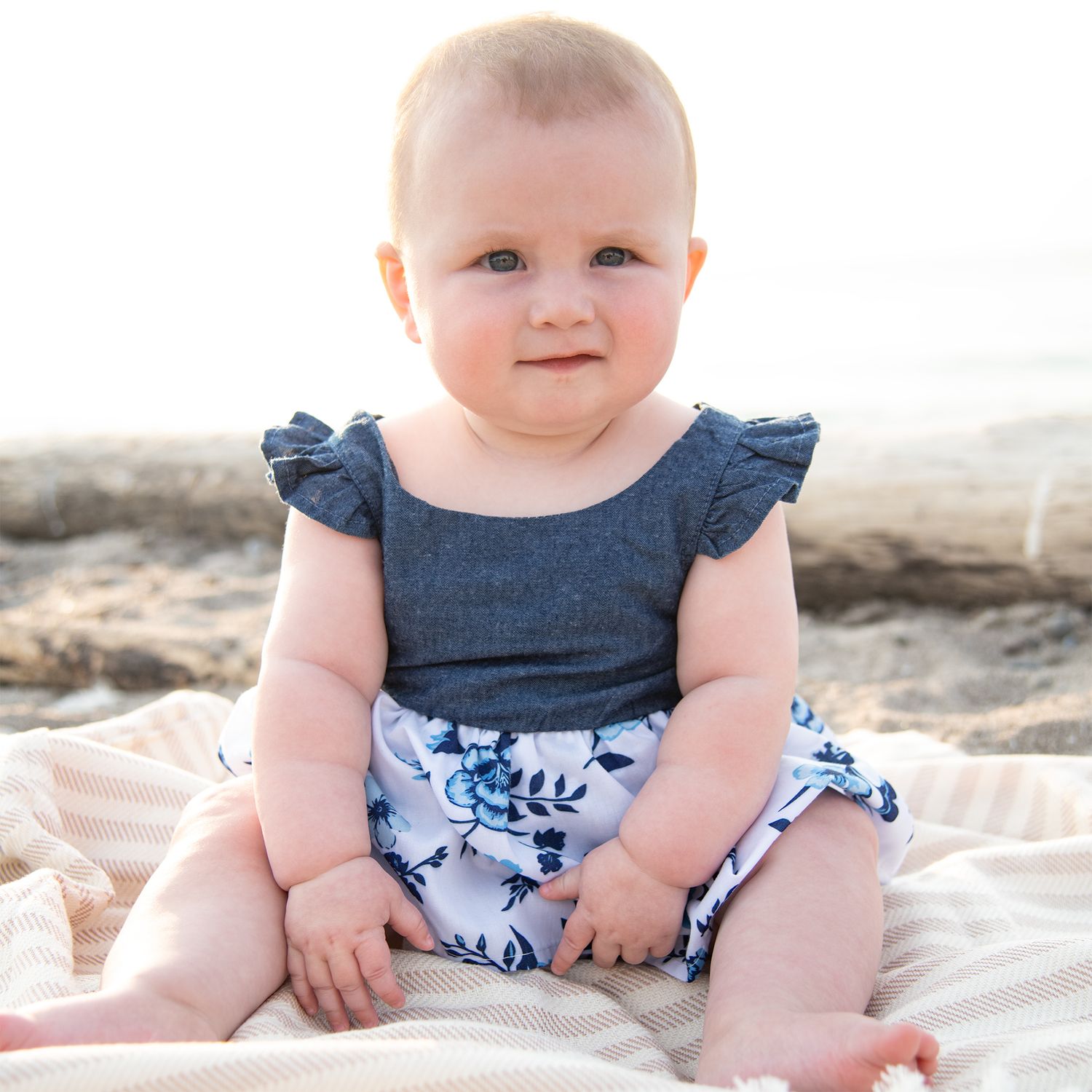 Small baby wearing a denim ruffle top and floral print bottoms sits on a light coloured blanket on the beach.