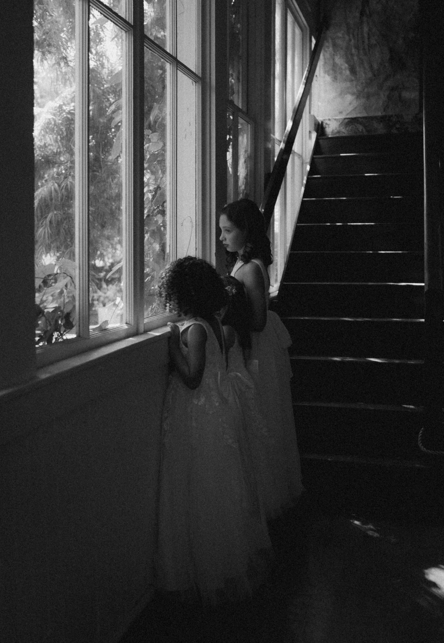 Two figures in white dresses stand by a window in a dark stairwell in a black and white photograph.