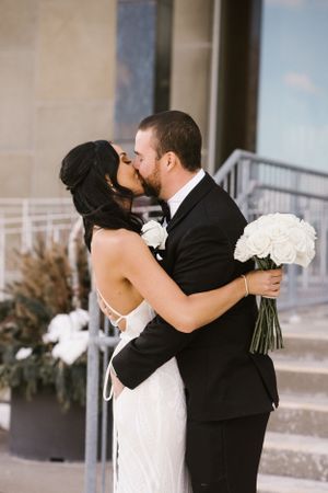 Romantic kiss between newlyweds outside venue with white bouquet.