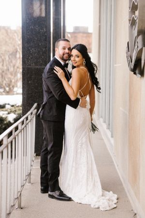 A romantic wedding couple poses on a balcony with the bride wearing a fitted lace gown and groom in classic black suit.