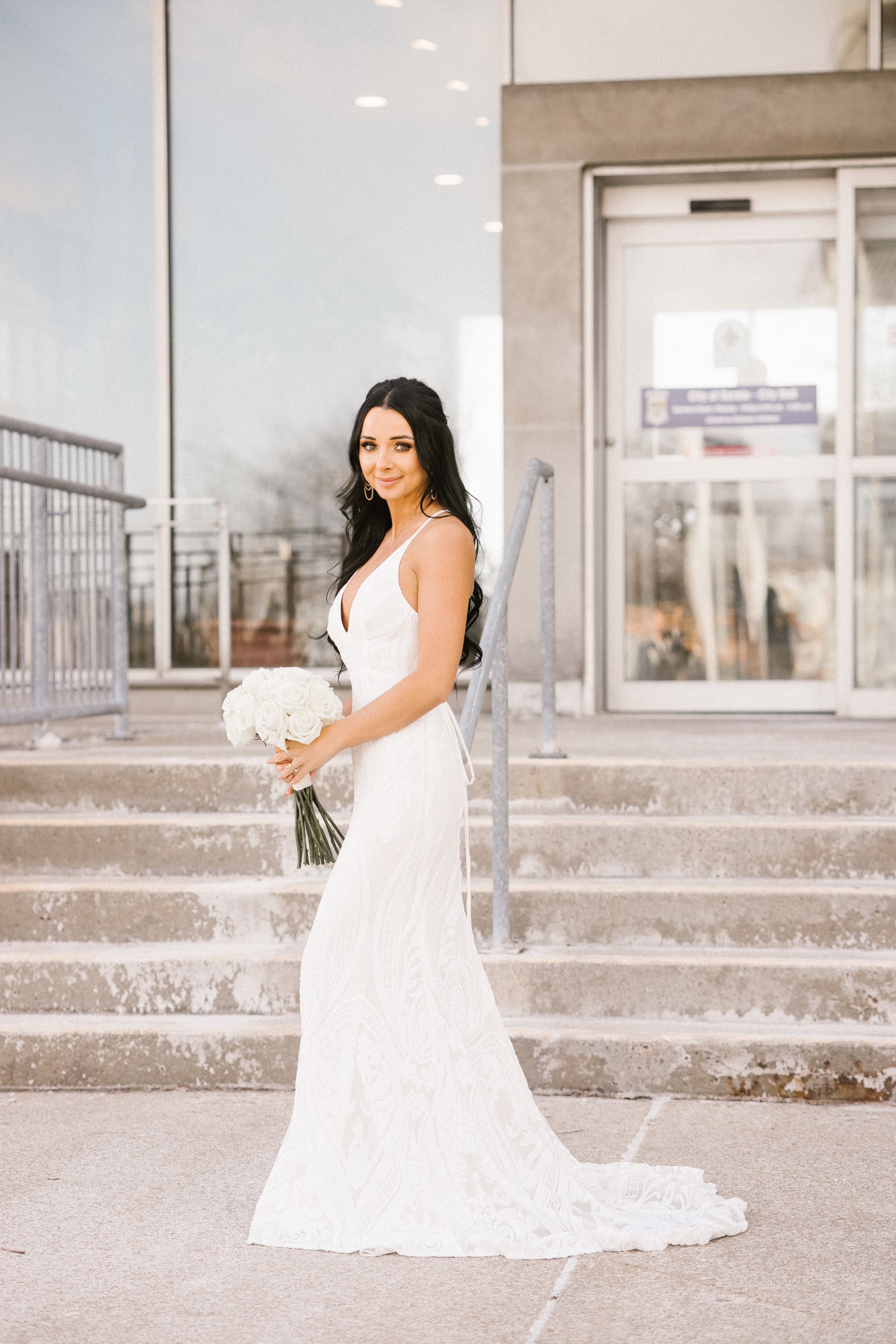 Bride in elegant white fitted dress holds bouquet while posing on outdoor concrete steps.