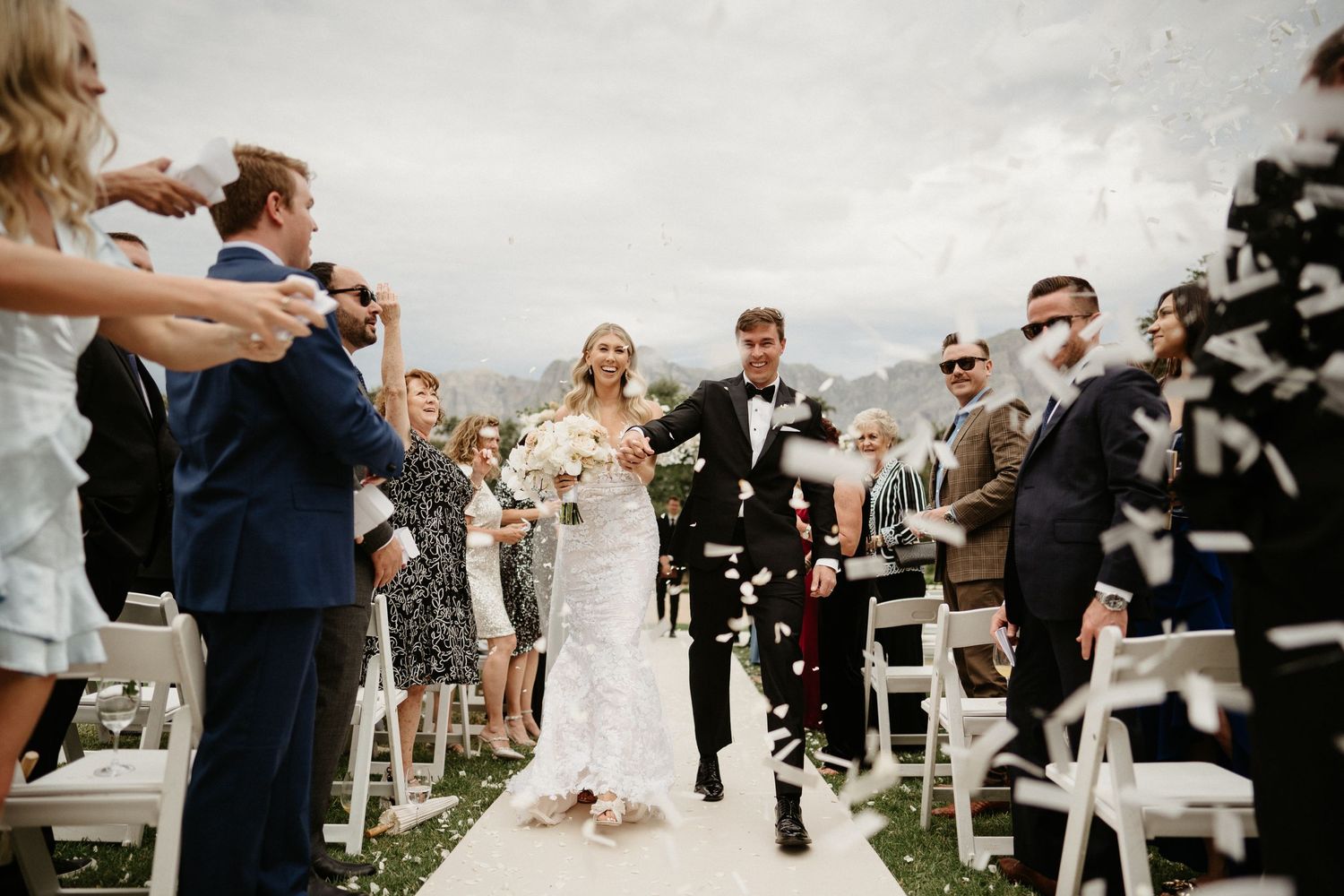 Newlyweds running down aisle while guests celebrate with confetti at outdoor wedding ceremony.