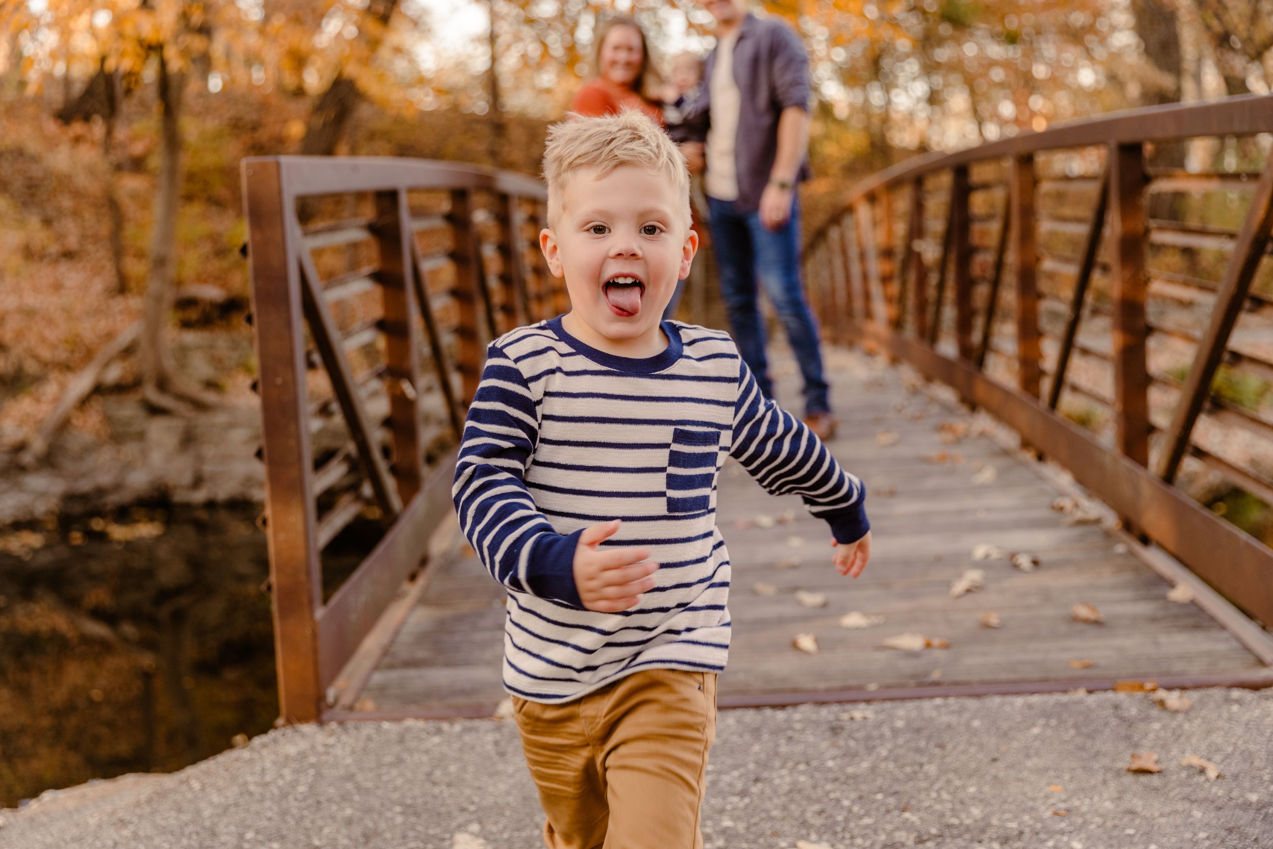 Young child walking and playing on wooden bridge pathway through autumn trees.