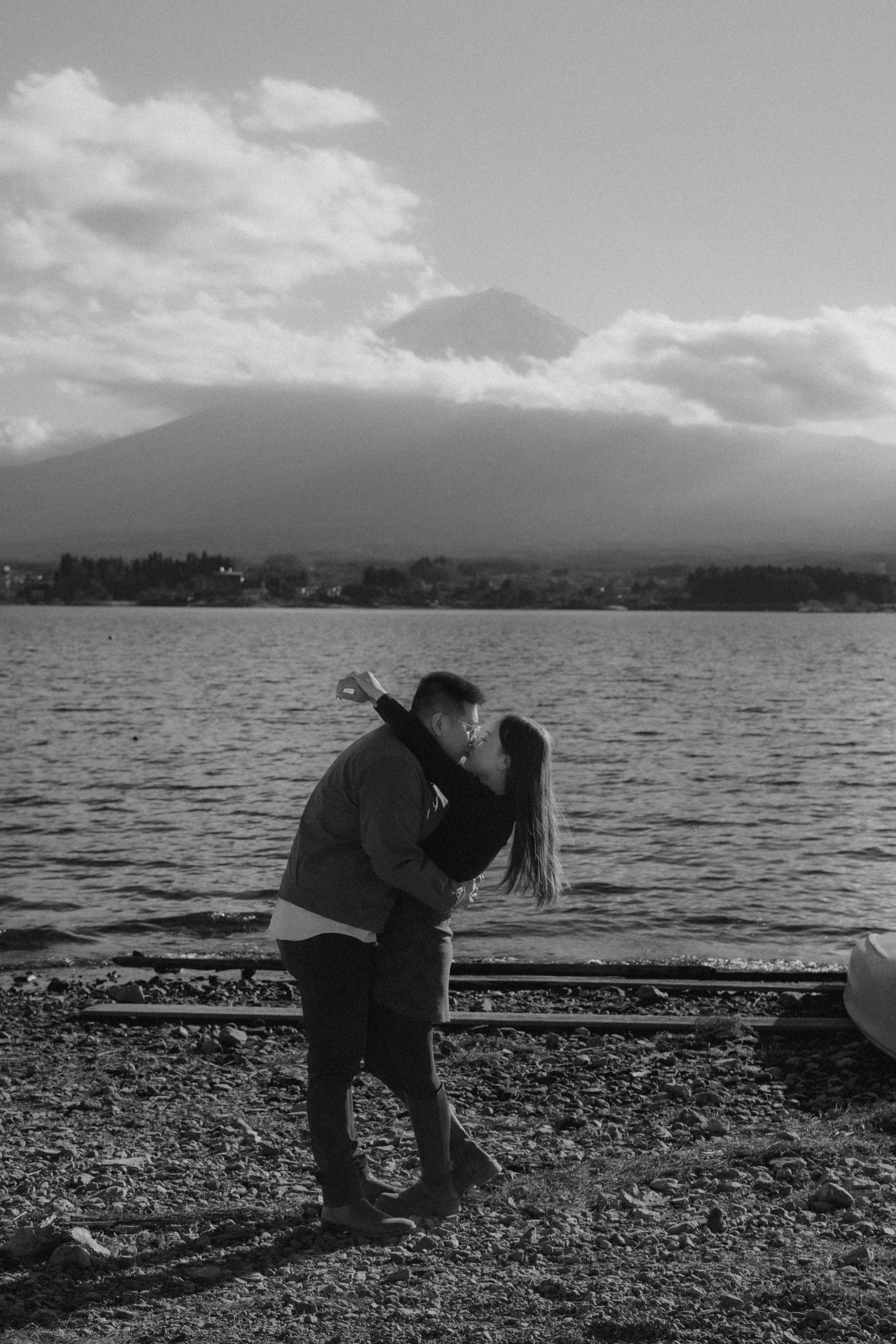 A silhouetted couple embraces on the rocky shore of a lake with Mount Fuji rising majestically in the cloudy background.