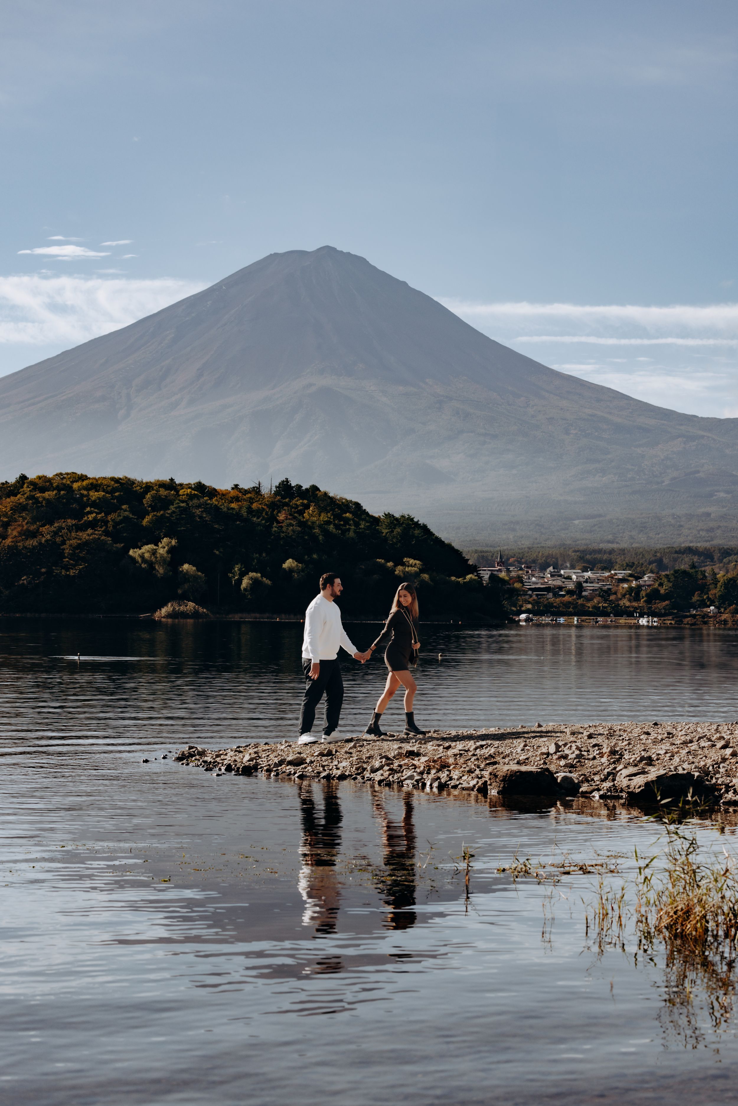 A couple standing on rocky shore with Mount Fuji reflecting in calm lake waters at sunset.