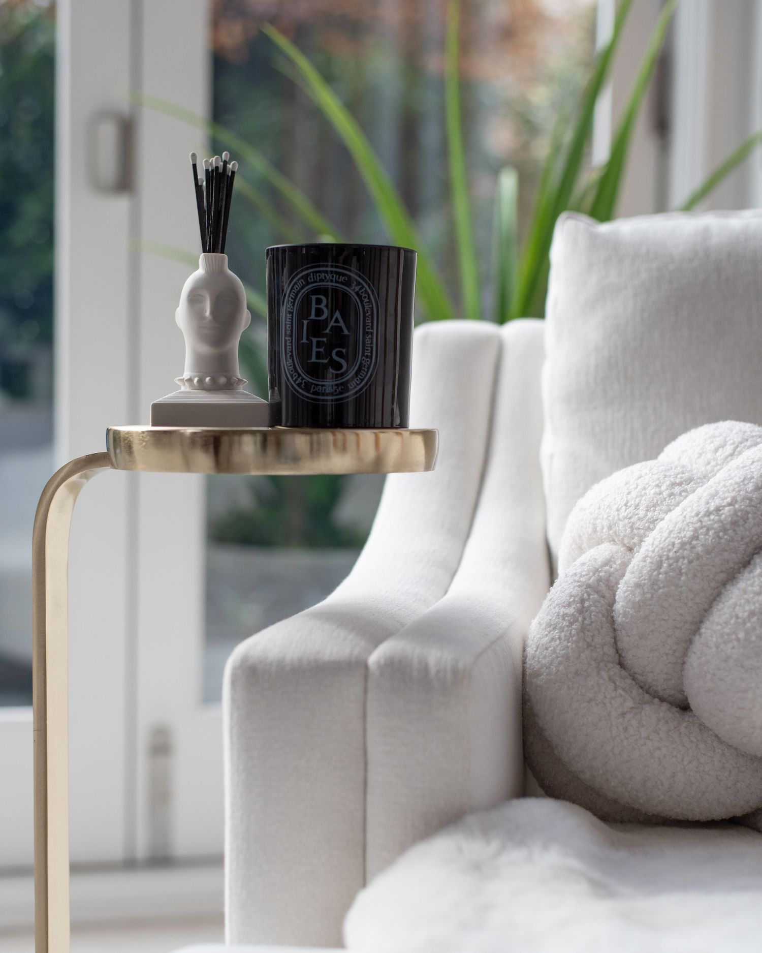 Luxury scented Diptyque candle and white diffuser on gold side table next to plush sofa with cozy throw pillows.