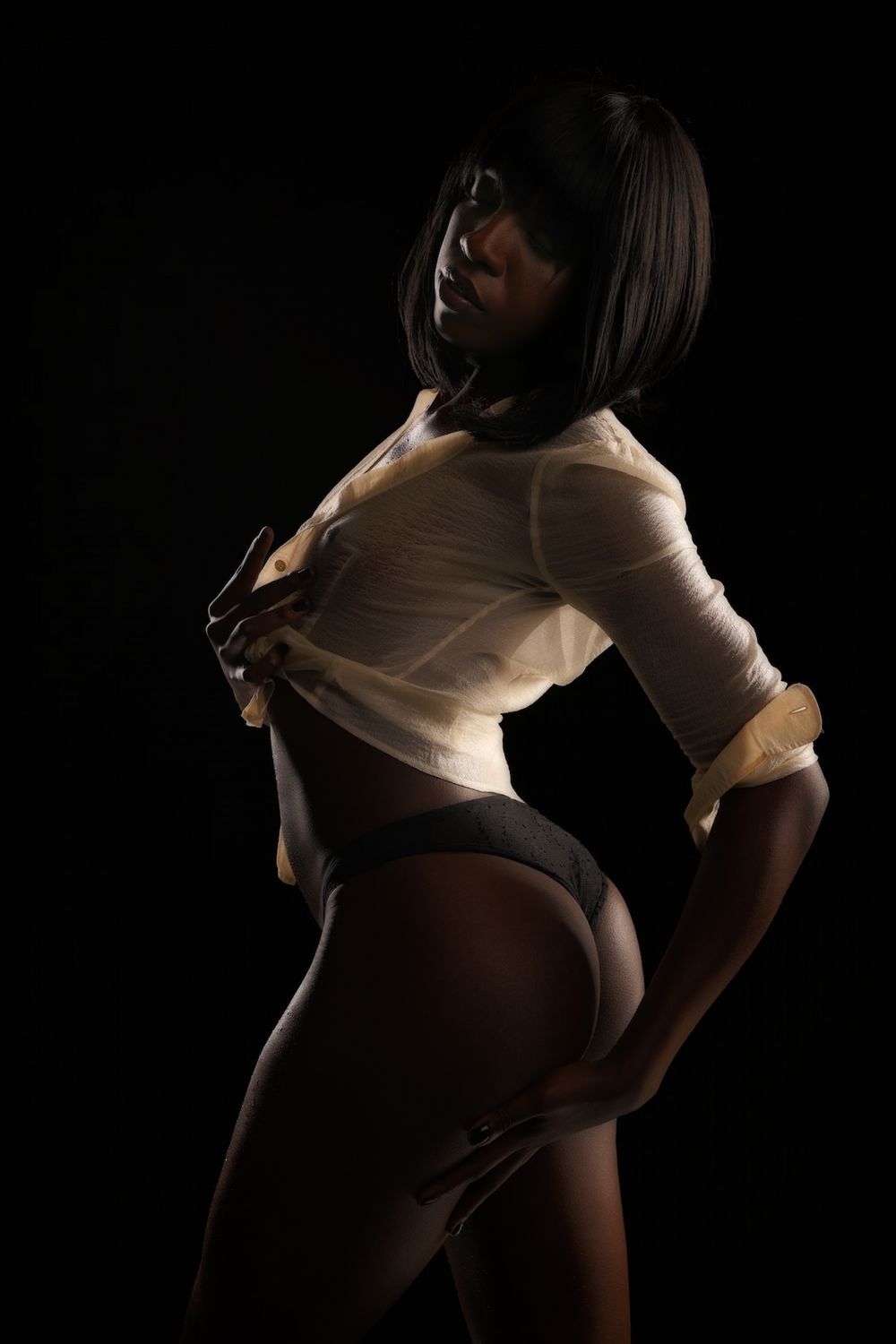 Moody low-key boudoir portrait in a sheer shirt and lingerie, with soft highlights on wet skin, a calm, confident pose emerging from shadow.