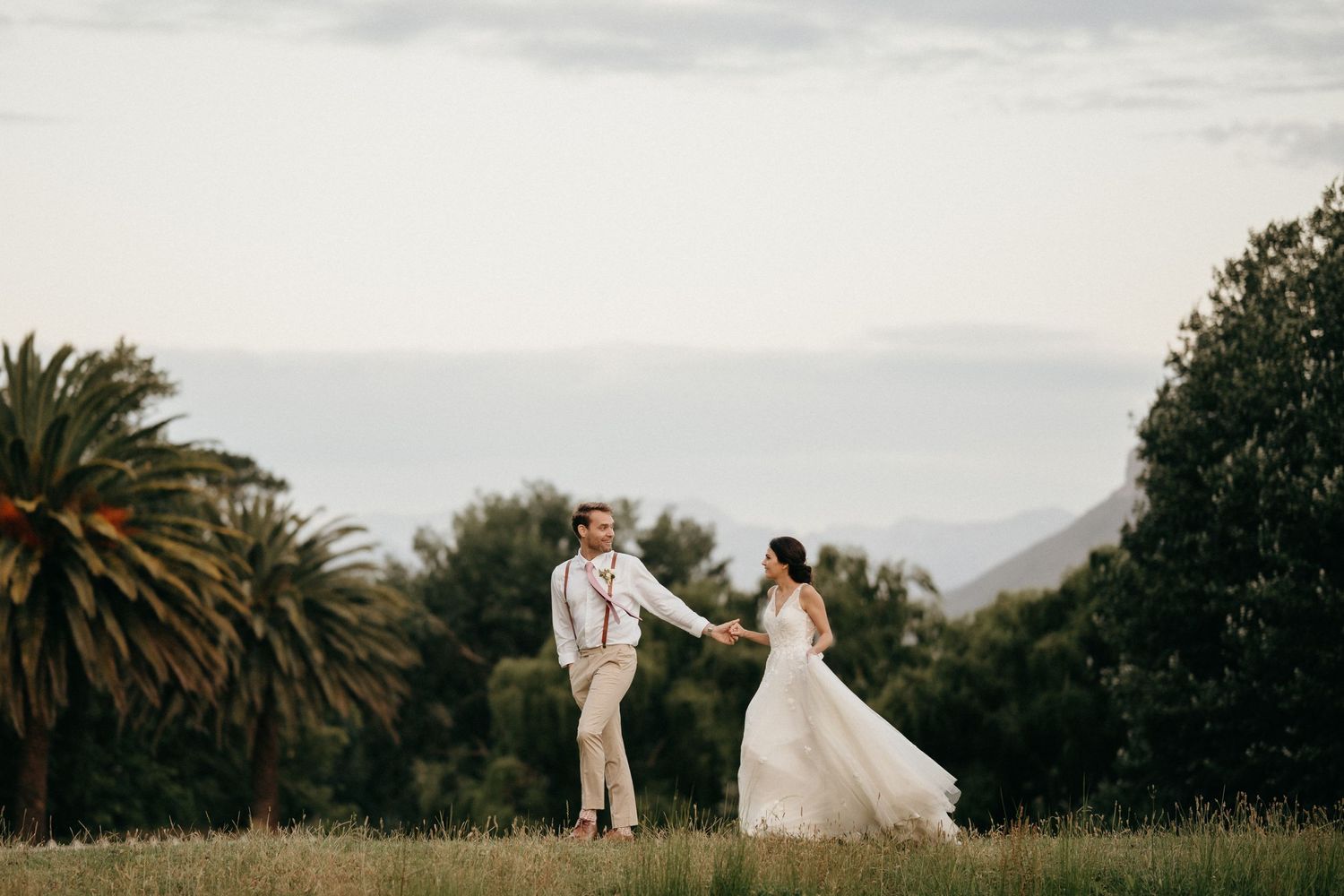 A couple shares a romantic moment in their wedding attire on a hilltop with palm trees and mountains in the background.