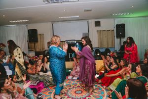 Guests in colorful traditional attire dance and celebrate on a decorative carpet during a festive indoor gathering.
