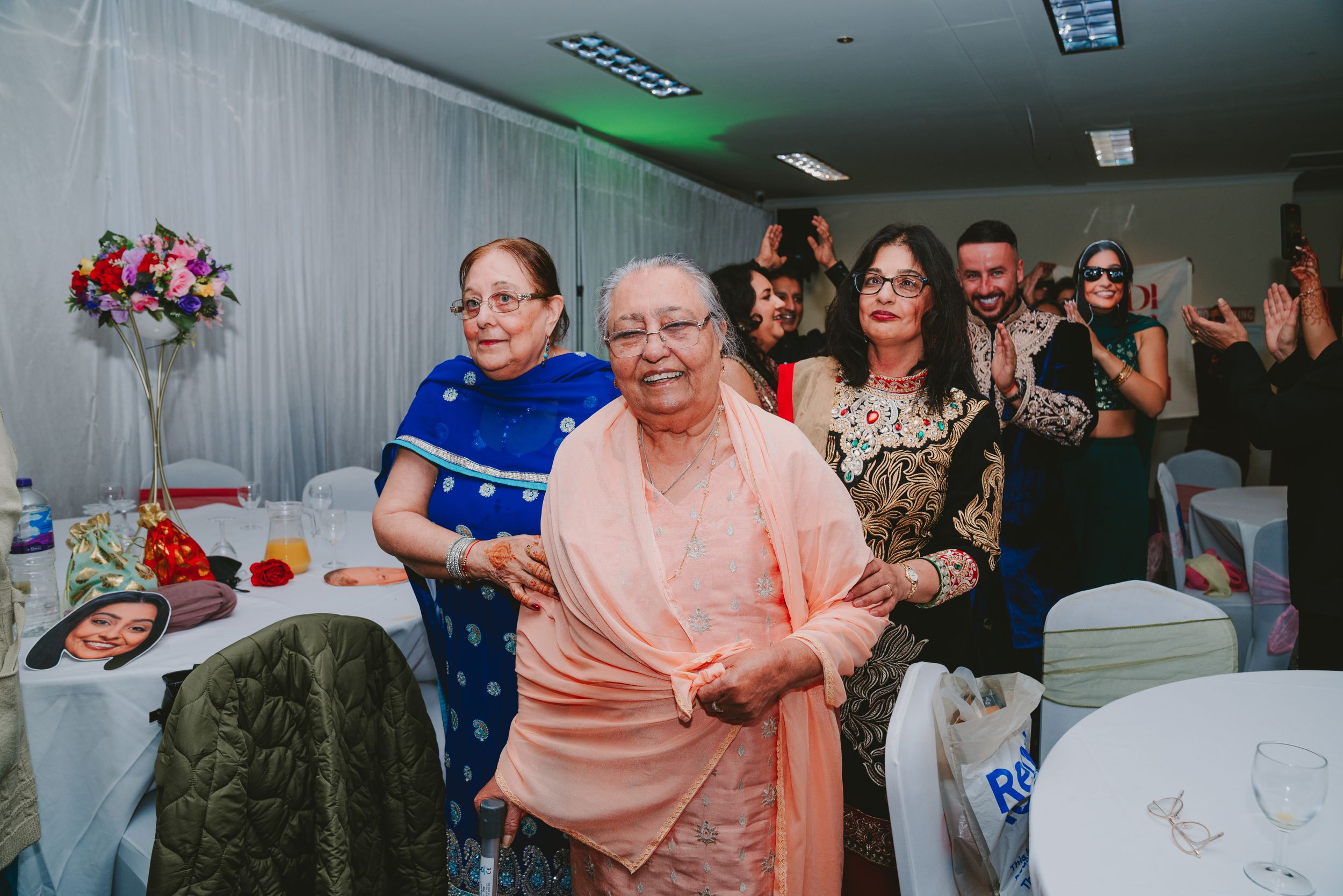 Party guests in formal attire gather at an event with floral decorations and white tablecloths.
