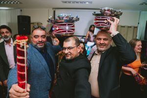 Three guests pose together while holding red and white striped ceremonial poles at an indoor celebration.