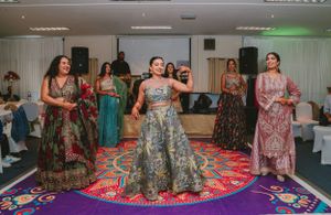 Dancers in colorful floral dresses performing on a decorative round carpet during an indoor celebration.