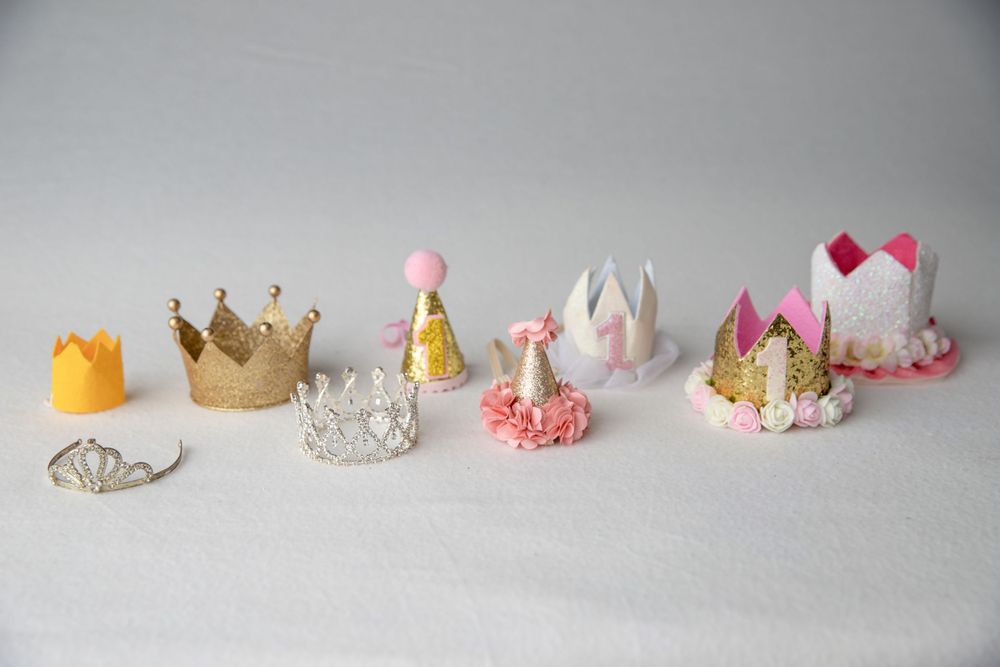Collection of decorative baby crowns and tiaras in gold, silver, pink, and white on gray background.