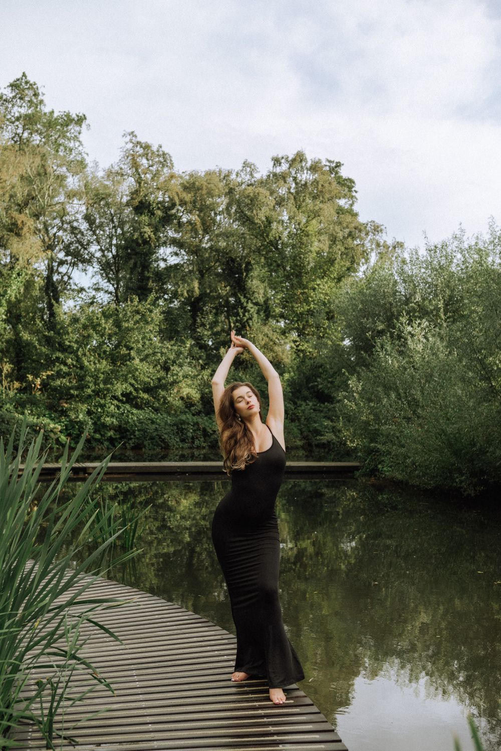 Expressive dance pose on a wooden dock by a tranquil pond surrounded by trees.