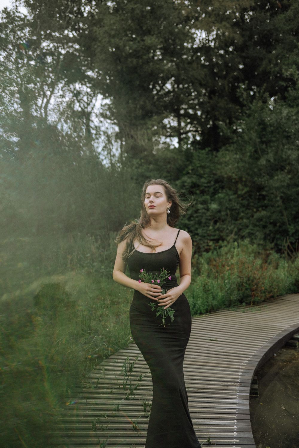 Elegant poses in a black dress against a serene waterfront landscape.