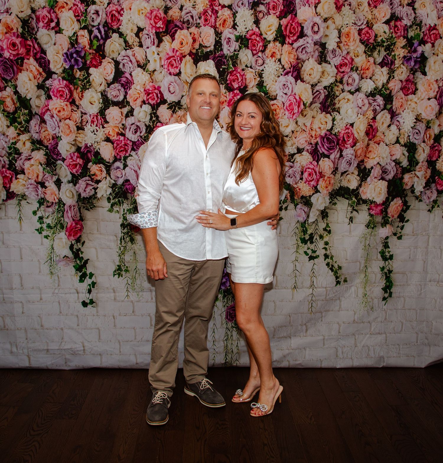 Couple posing against floral wall backdrop with pink and white roses.