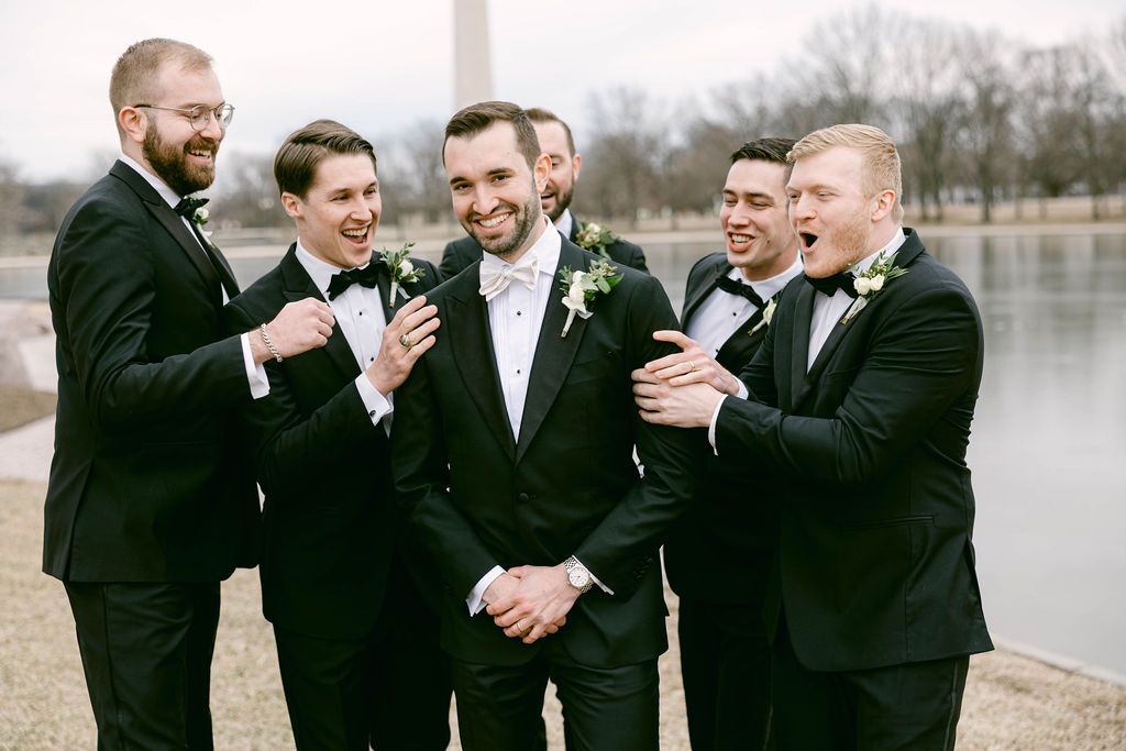 A group wearing black suits laughs together during a wedding photo session by a lake.