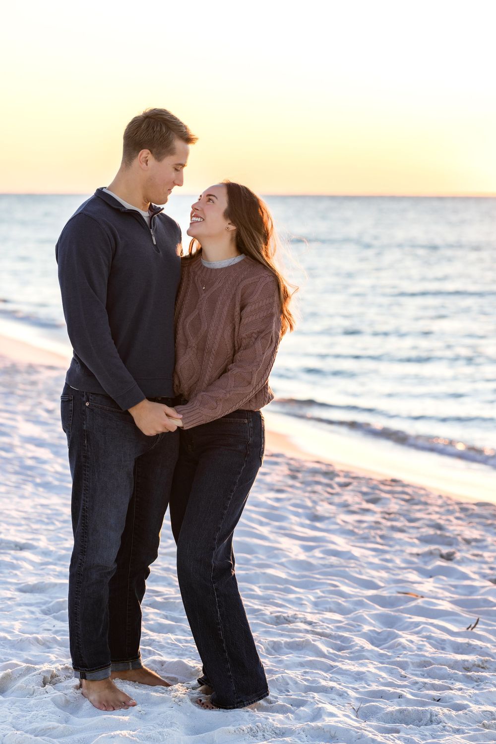 A couple shares tender moments on a sandy beach at sunset with waves in the background.