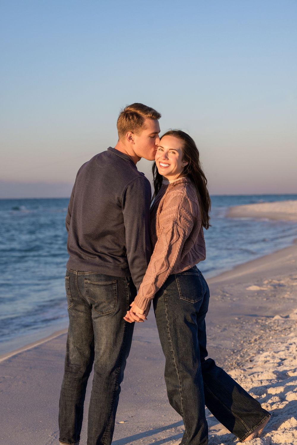 Couple embracing on a sandy beach at sunset with ocean waves in the background.