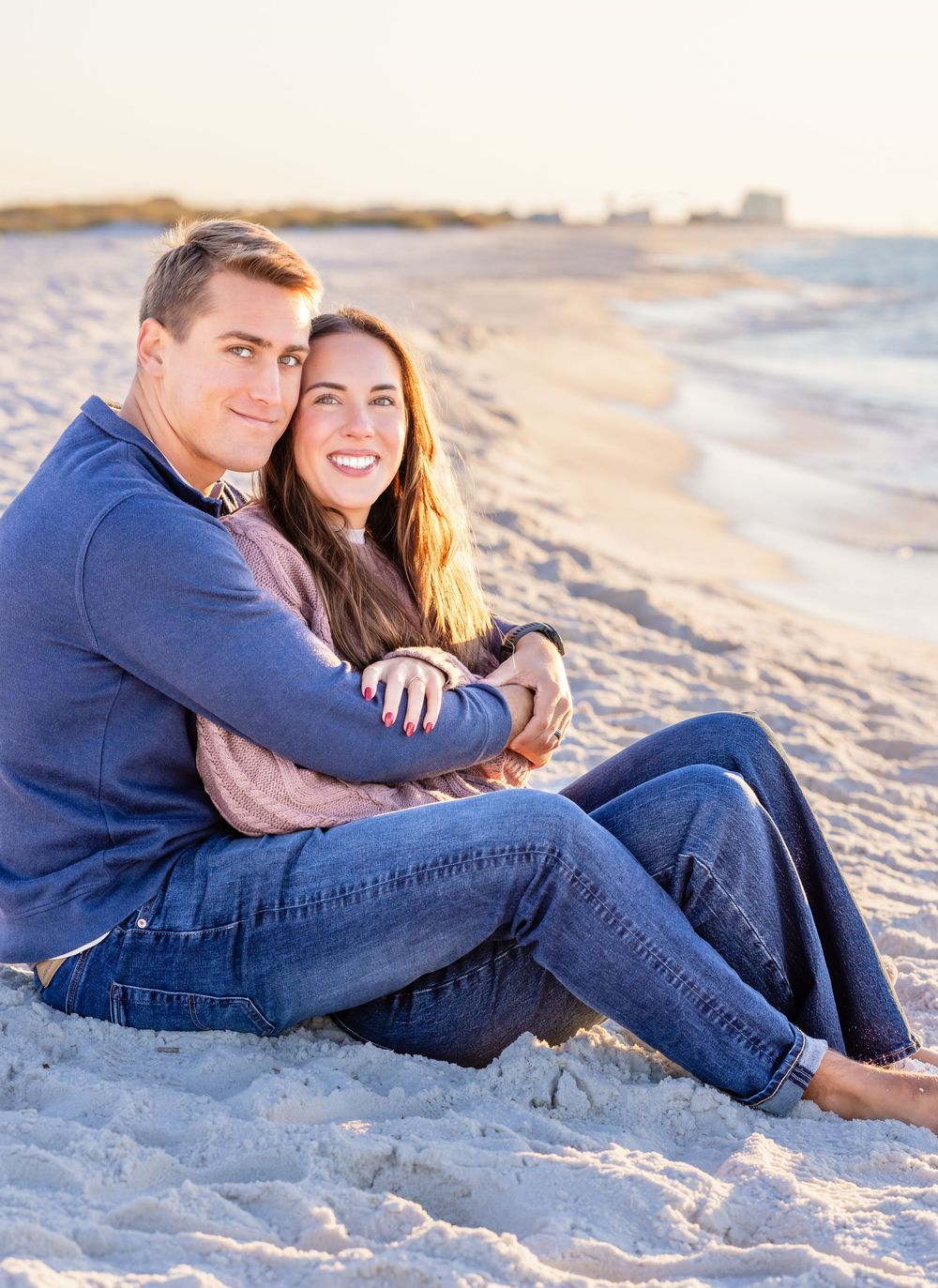 A couple sits together on a sandy beach at sunset enjoying a romantic moment in casual denim attire.