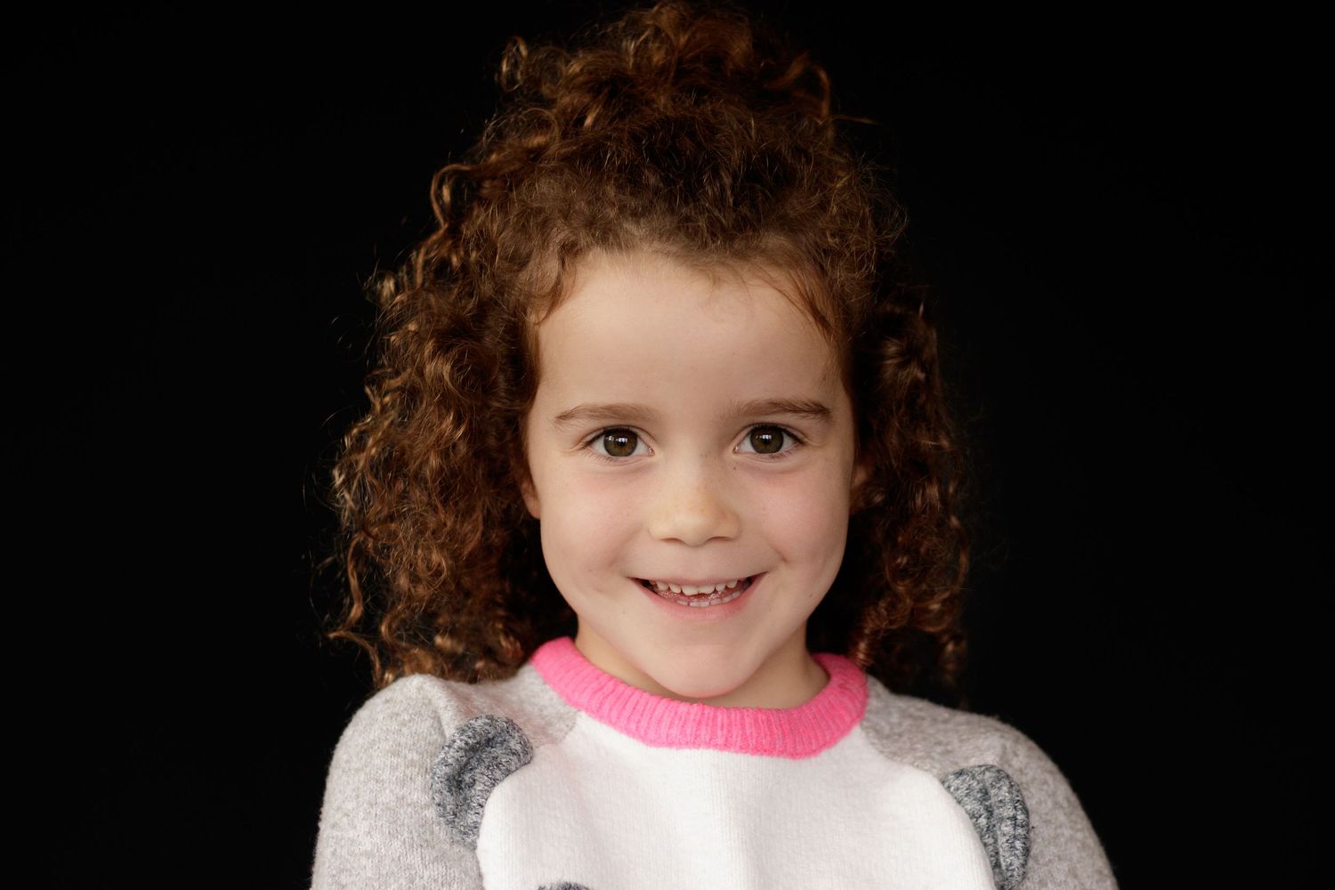 Smiling young girl with curly auburn hair wearing a pink and white graphic shirt against a dark background.