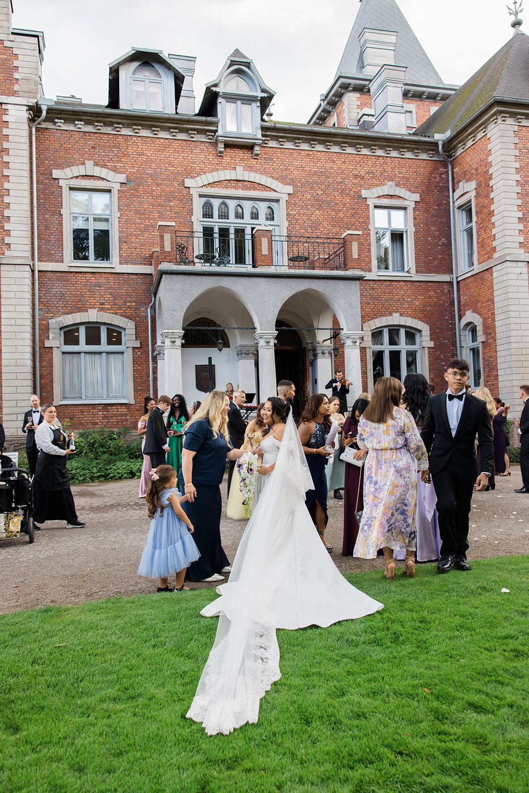 Wedding guests gather on the lawn outside a grand brick mansion with ornate architectural details during an elegant celebration.