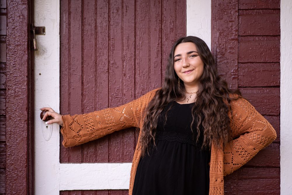 Person in black dress and brown cardigan poses against rustic red and white door.Middletown NJ