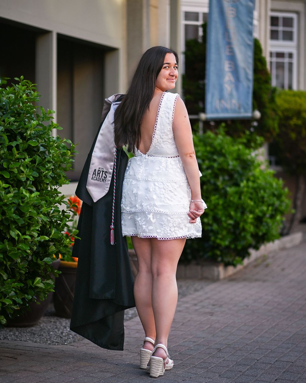 Young woman in white lace mini dress holding graduation gown outdoors, celebrating college graduation.