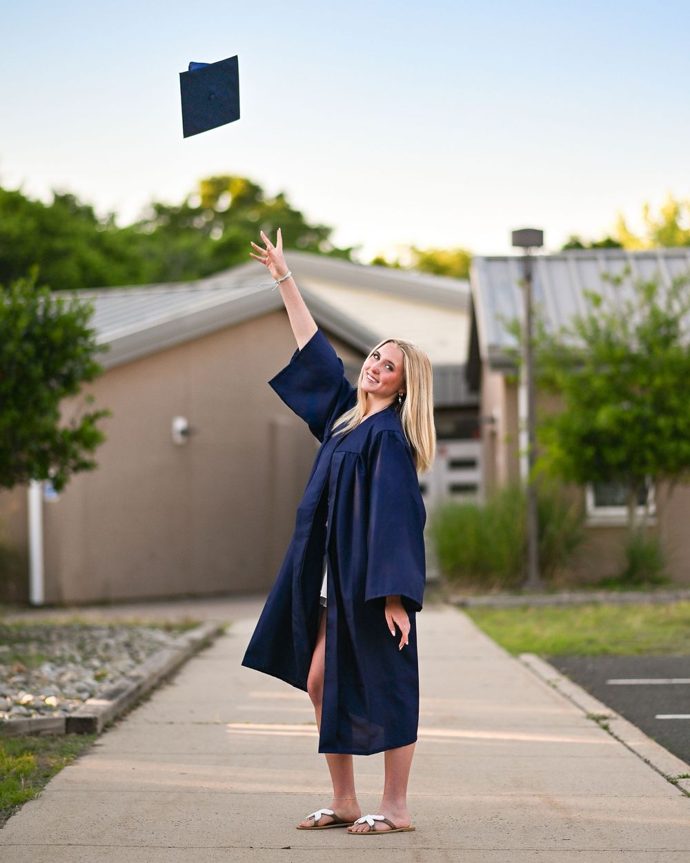 Smiling graduate in navy blue cap and gown tossing mortarboard in air on sunny day outdoors.