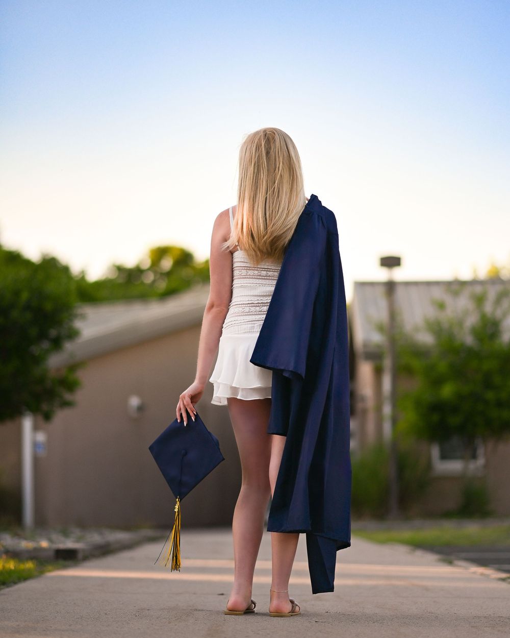 Blonde graduate holding navy cap and gown outdoors at sunset, standing barefoot on a pathway.