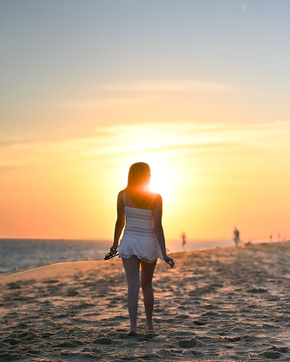 Woman in white dress standing on sandy beach at golden sunset, silhouetted against vibrant orange sky.