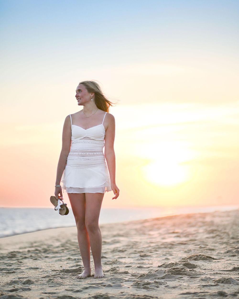 Young woman in white dress walking barefoot on sandy beach at sunset, holding sandals with hair blowing in breeze.