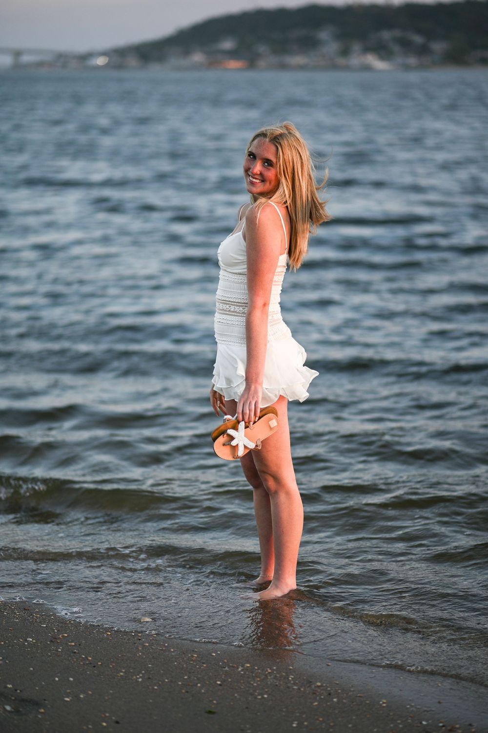 Smiling blonde woman in white dress standing barefoot in ocean waves at beach during sunset.