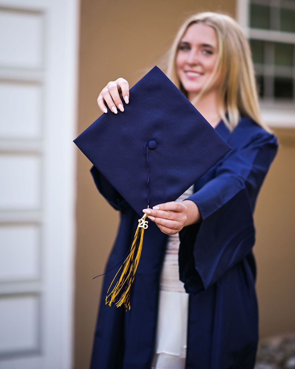 Smiling blonde woman in navy blue graduation gown holding mortarboard cap with gold tassel outdoors.