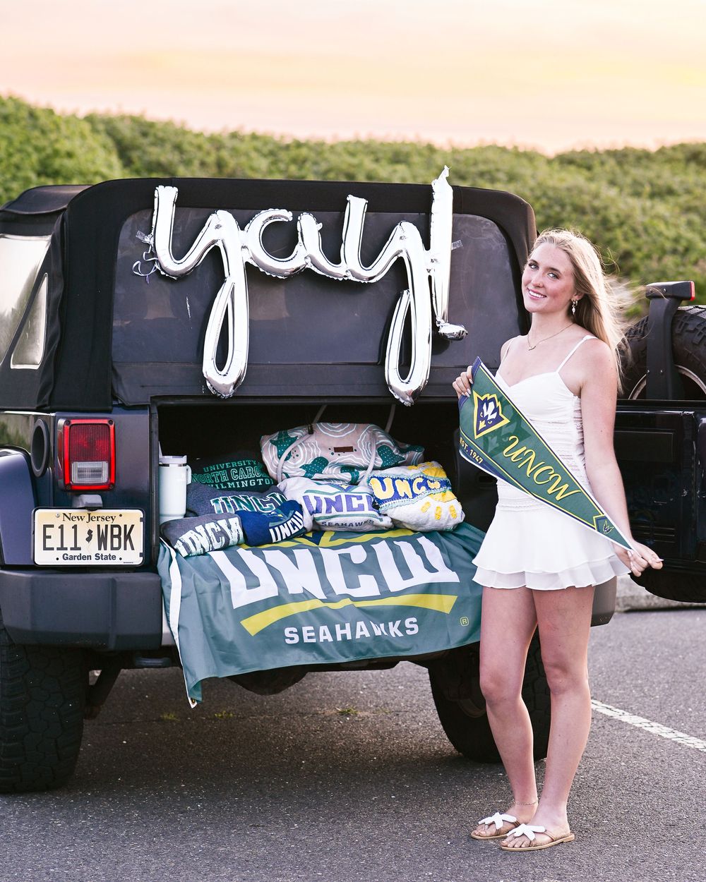 Young woman in white dress holding UNCW Seahawks pennant beside decorated Jeep with yay balloon for college acceptance celebration.