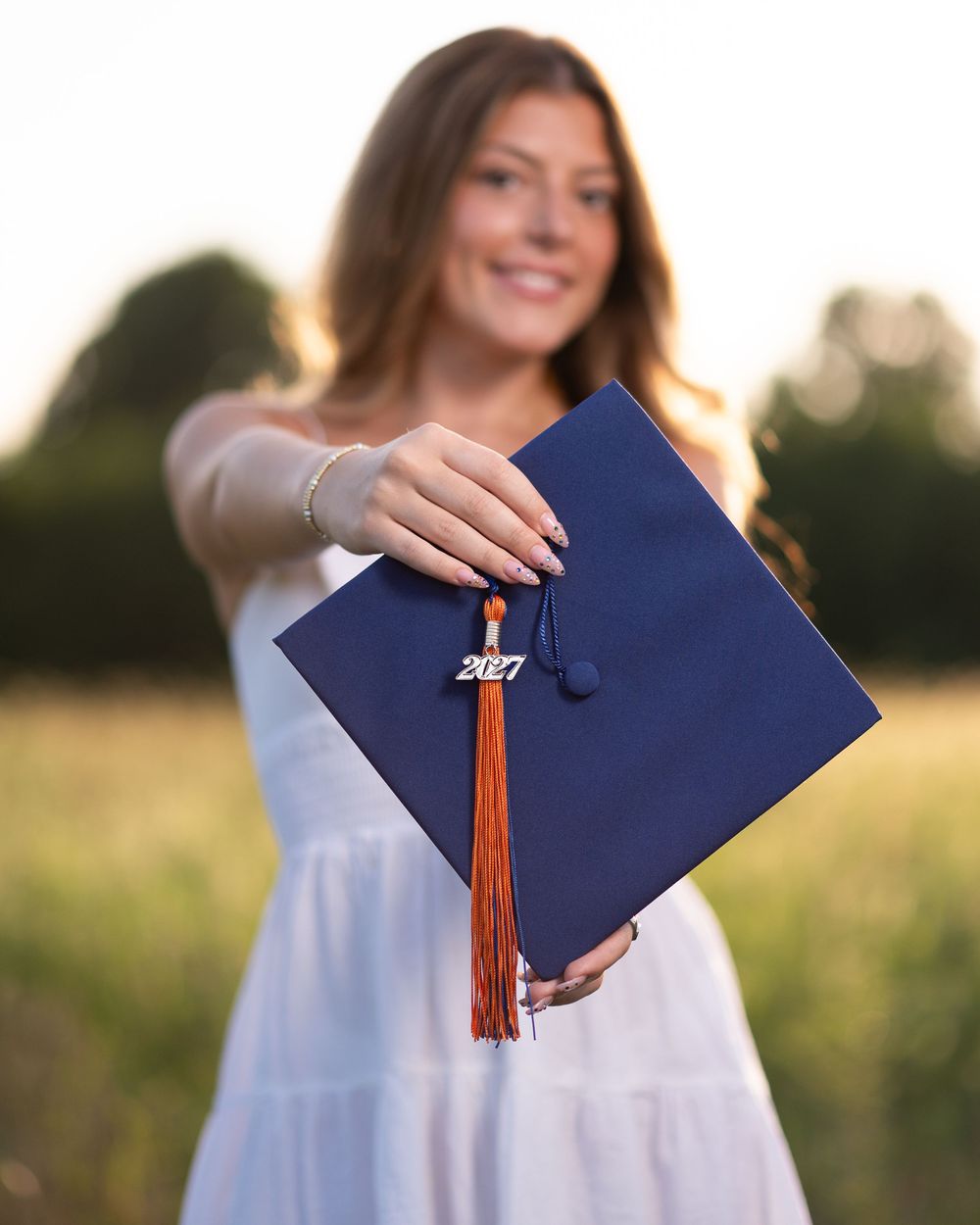 Young woman in white dress holding a navy blue 2027 graduation cap with orange tassel outdoors at golden hour.