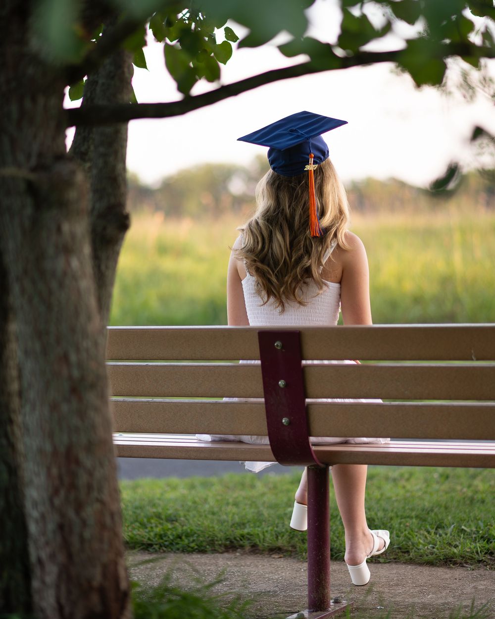 A graduate in a blue cap sits on a park bench facing a green meadow, long blonde hair cascading down her back.