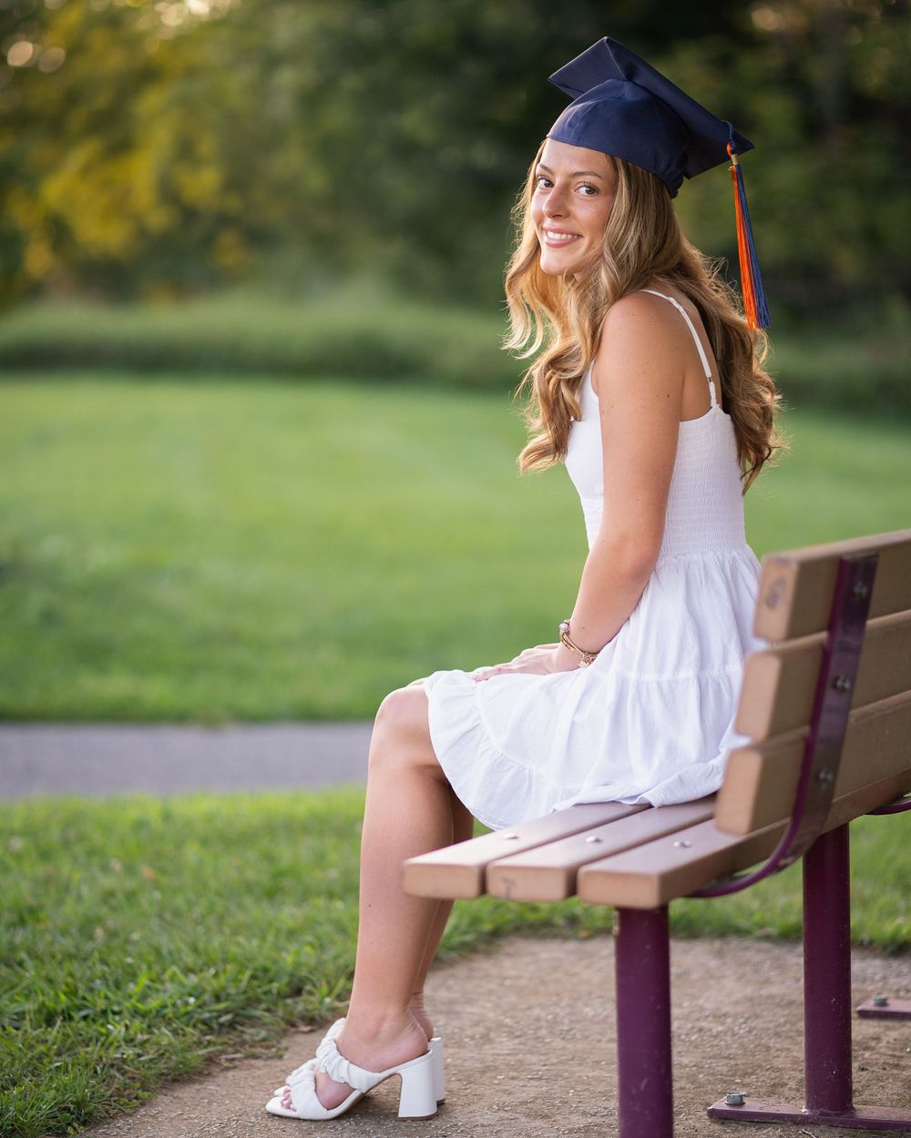 Young woman in white dress and navy graduation cap sits smiling on a park bench outdoors.