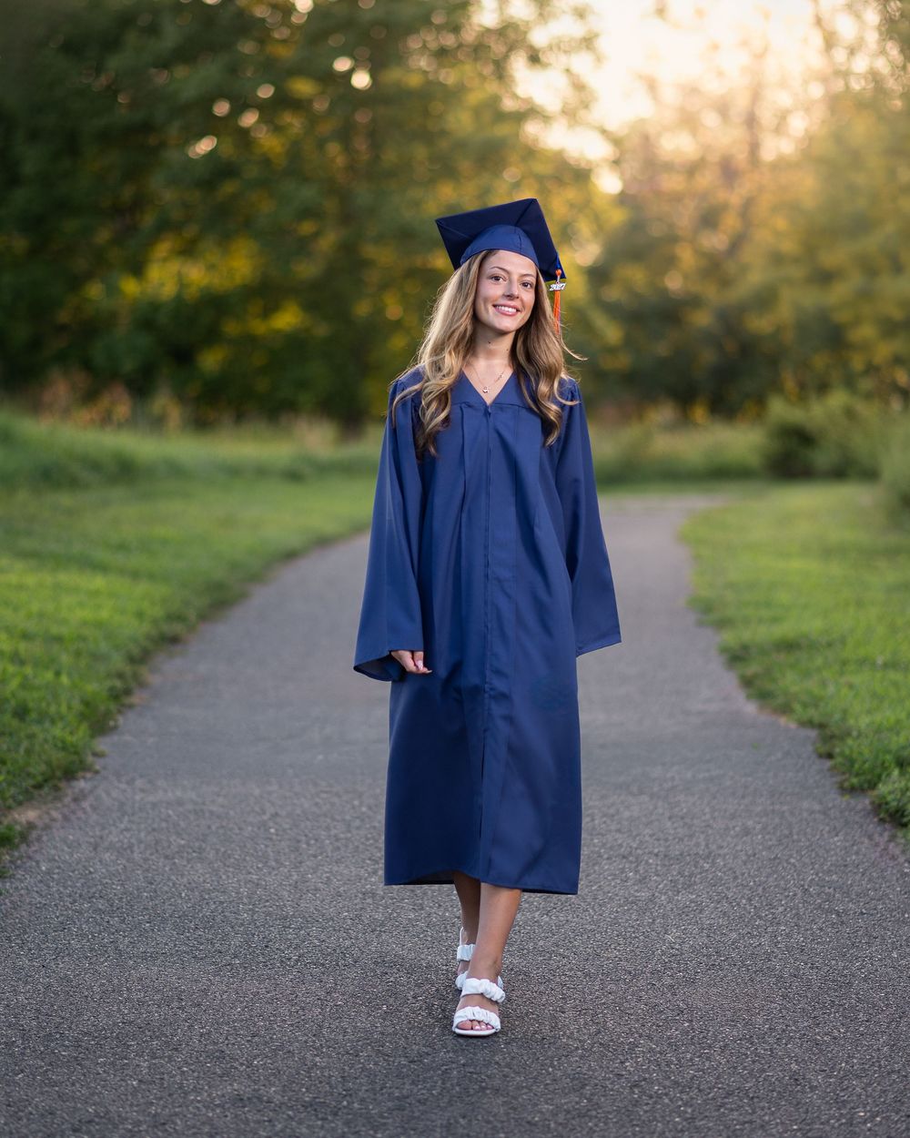 Smiling graduate in navy blue cap and gown walking on a tree-lined path during golden hour.