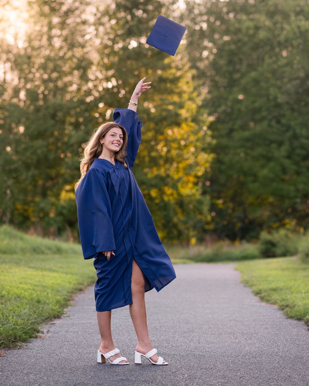 A smiling graduate in navy blue cap and gown tosses her mortarboard in the air on a tree-lined path.