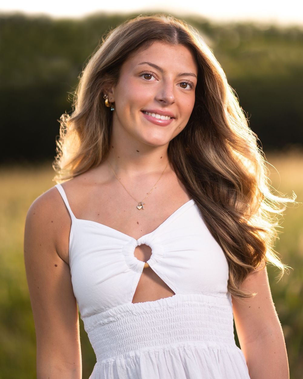 Young woman with long brown hair smiling outdoors in a white sundress during golden hour.