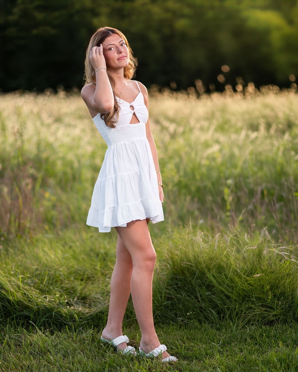 Young woman in white mini dress posing in a sunlit green field during golden hour photography session.