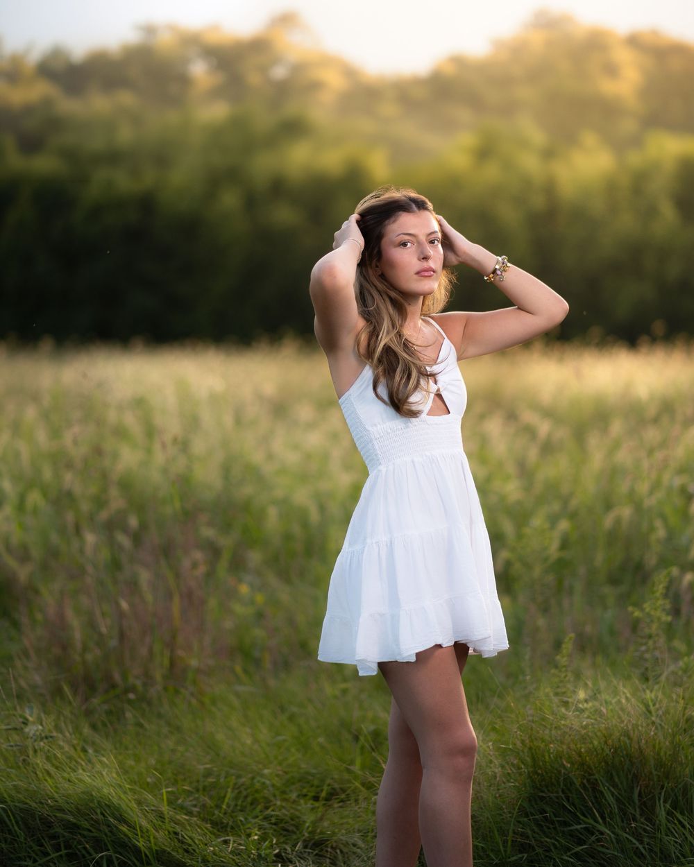 Young woman in white sundress poses gracefully in a sunlit meadow with lush green trees in the background.