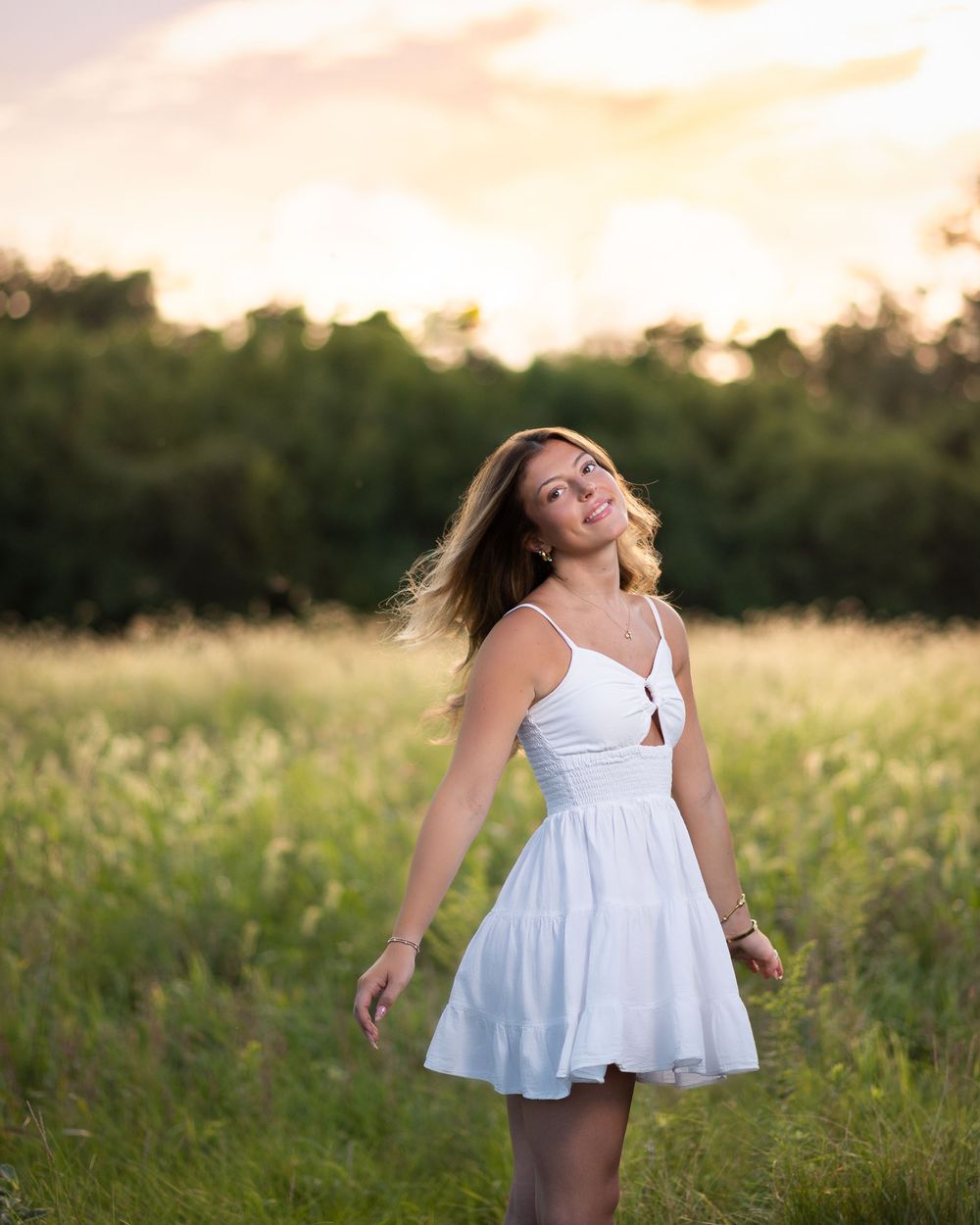 Young woman in white sundress twirling in a green meadow at golden hour sunset.