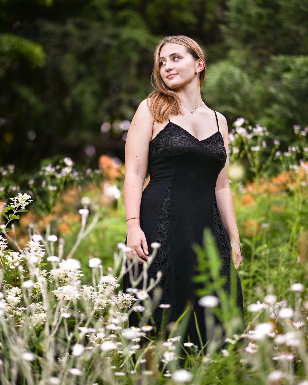 Young woman in black lace dress standing among wildflowers in a lush garden, gazing upward serenely.