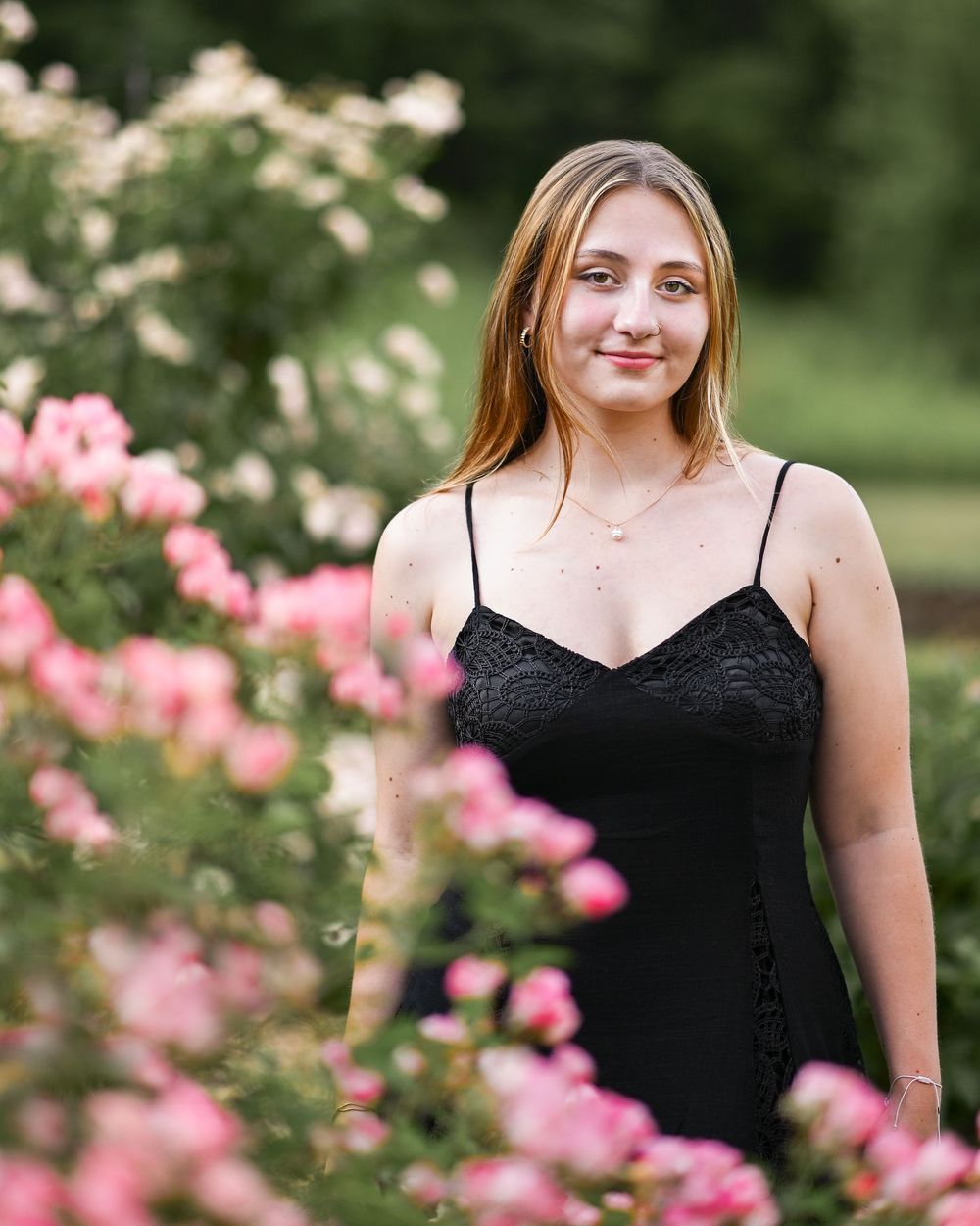 Young woman in black lace dress posing among pink and white roses in a lush garden setting.